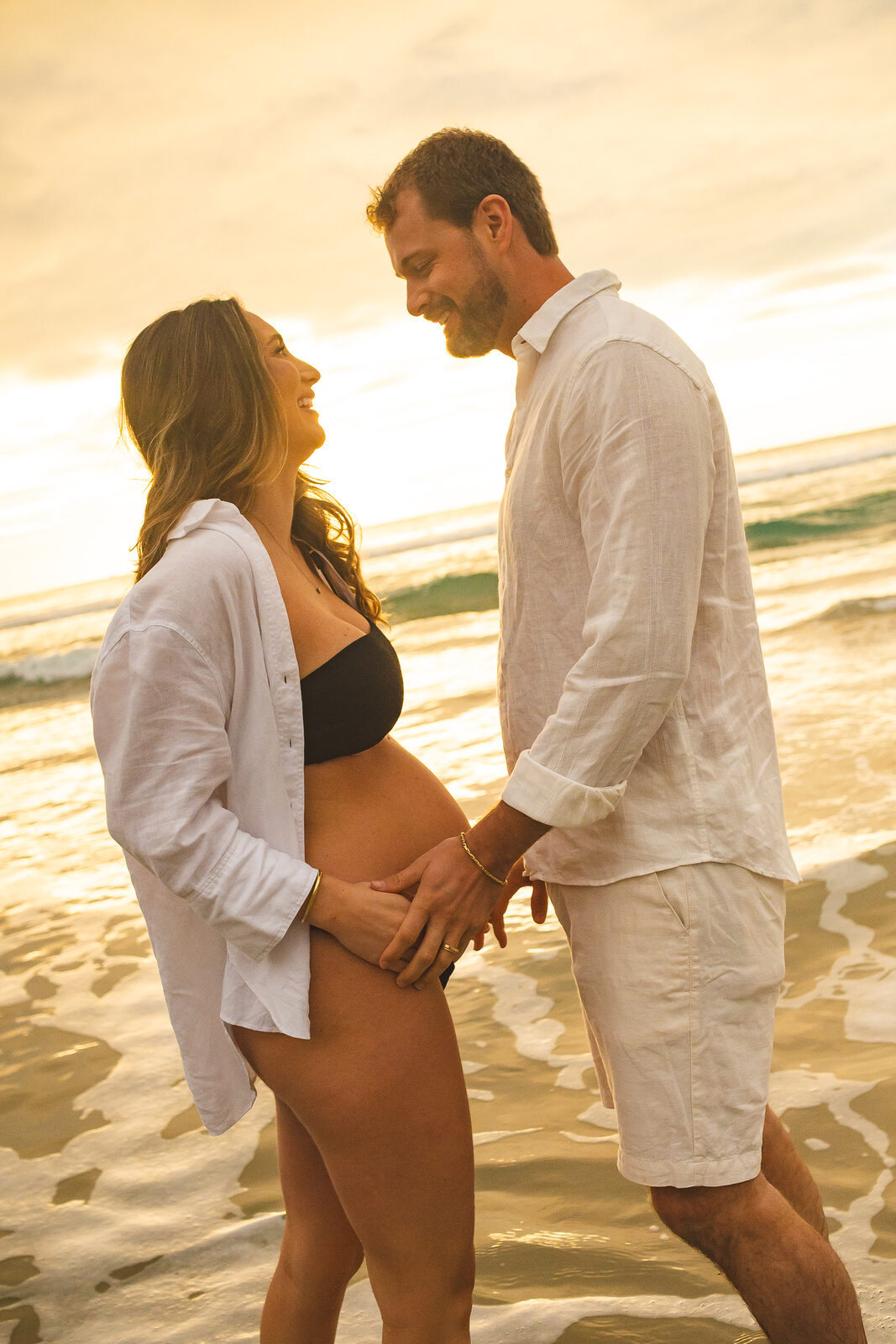 Casal na areia na luz dourada, as mãos sobre a barriga