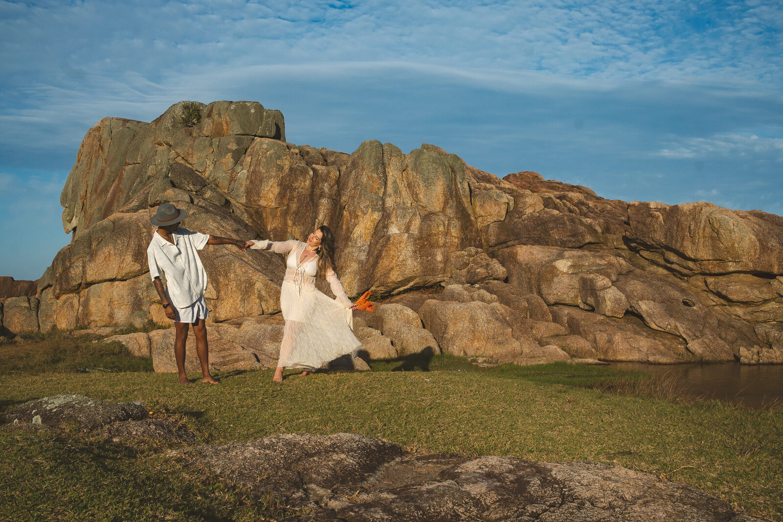 Casal ao pé de um costão de pedra, campo verde e mar ao fundo